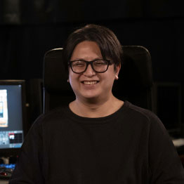 A smiling person wearing glasses sits in a dark room with computer monitors in the background, possibly a studio or editing suite, on a ZEISS professional product website.