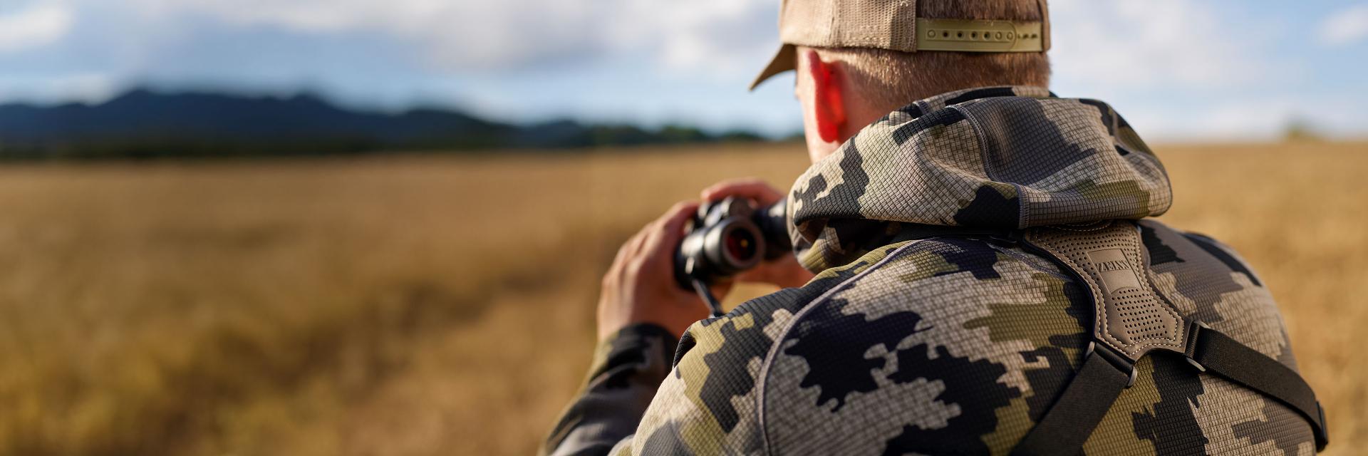 A person in camouflage clothing is using ZEISS binoculars while observing a vast open field with mountains in the background.