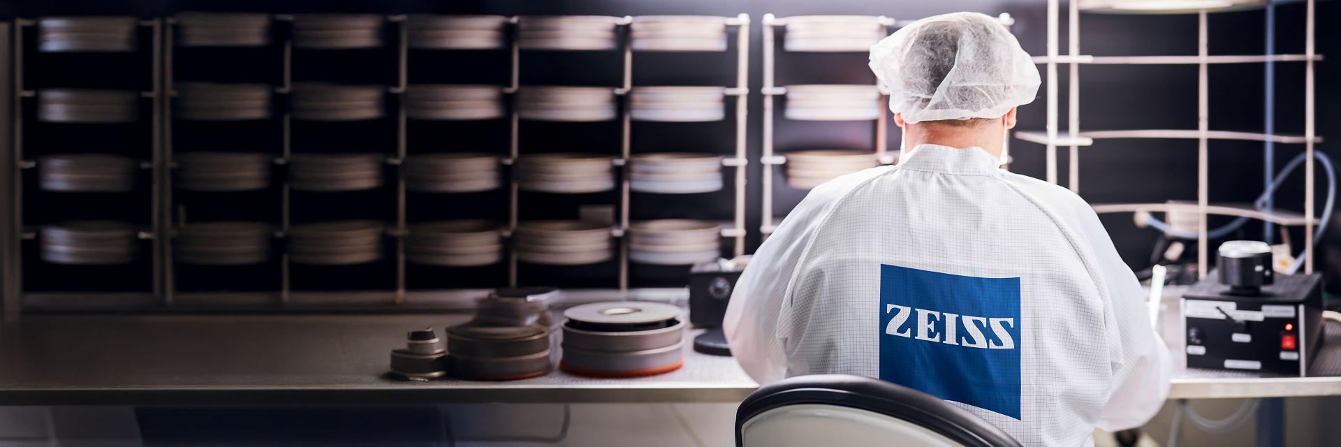 A technician in a cleanroom environment wearing a ZEISS lab coat is working at a workstation surrounded by stacked optical components.
