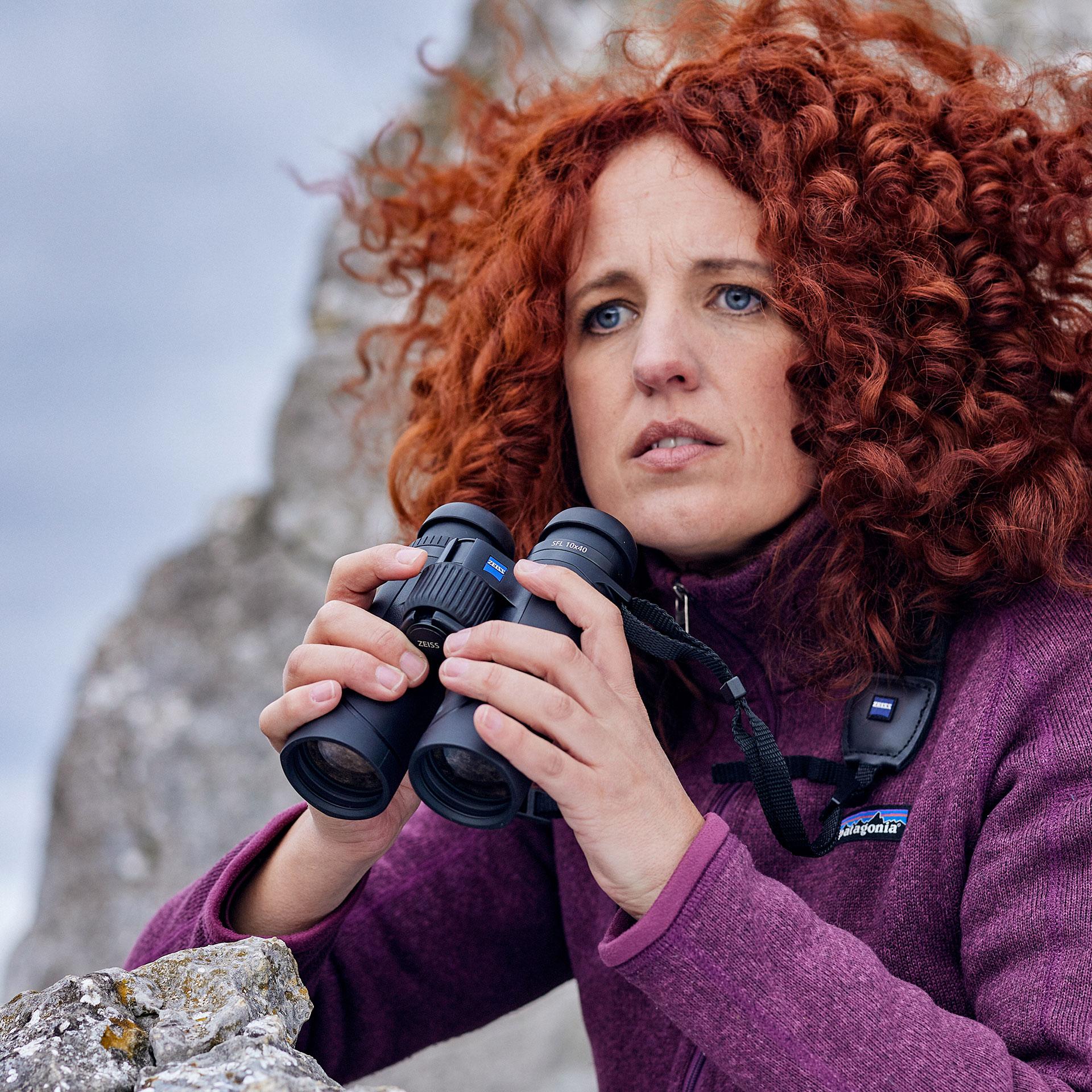 A woman with curly red hair, dressed in a purple jacket, holds a pair of ZEISS binoculars while observing from a rocky outdoor setting.