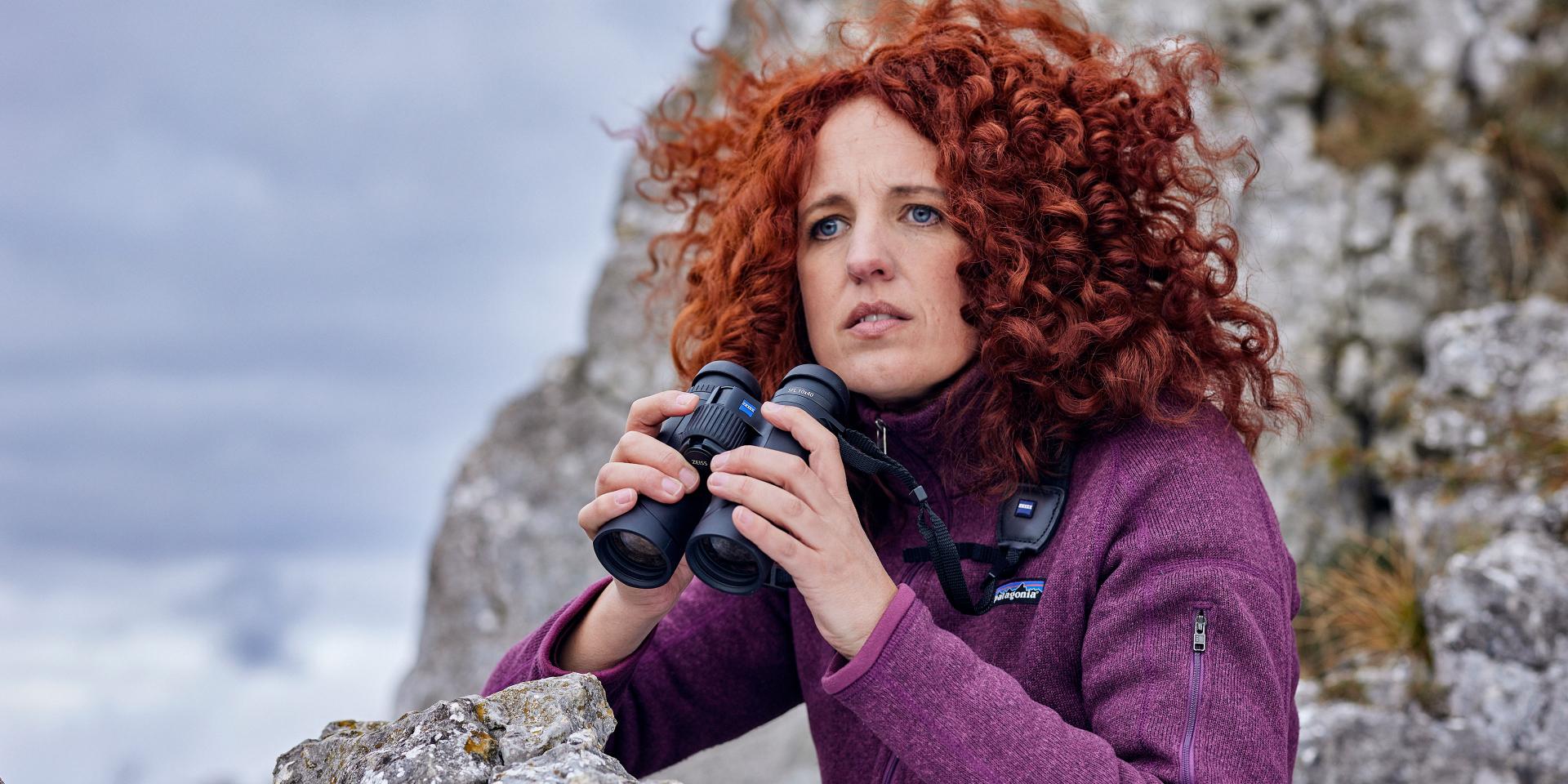A woman with curly red hair, dressed in a purple jacket, holds a pair of ZEISS binoculars while observing from a rocky outdoor setting.