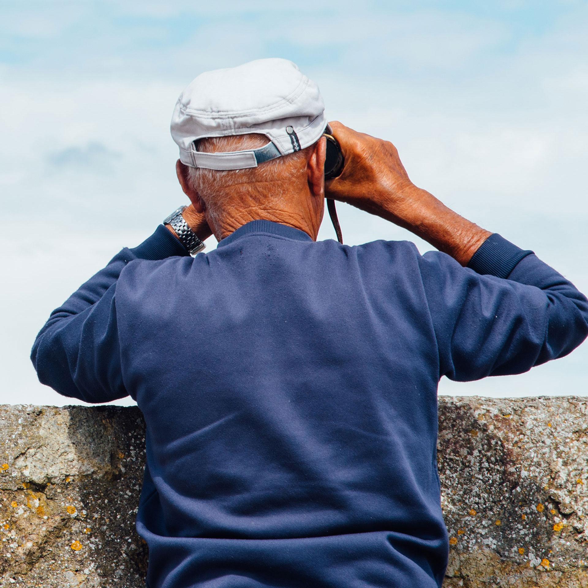 An older man wearing a blue sweater and white cap looks through binoculars over a stone wall toward the sky.