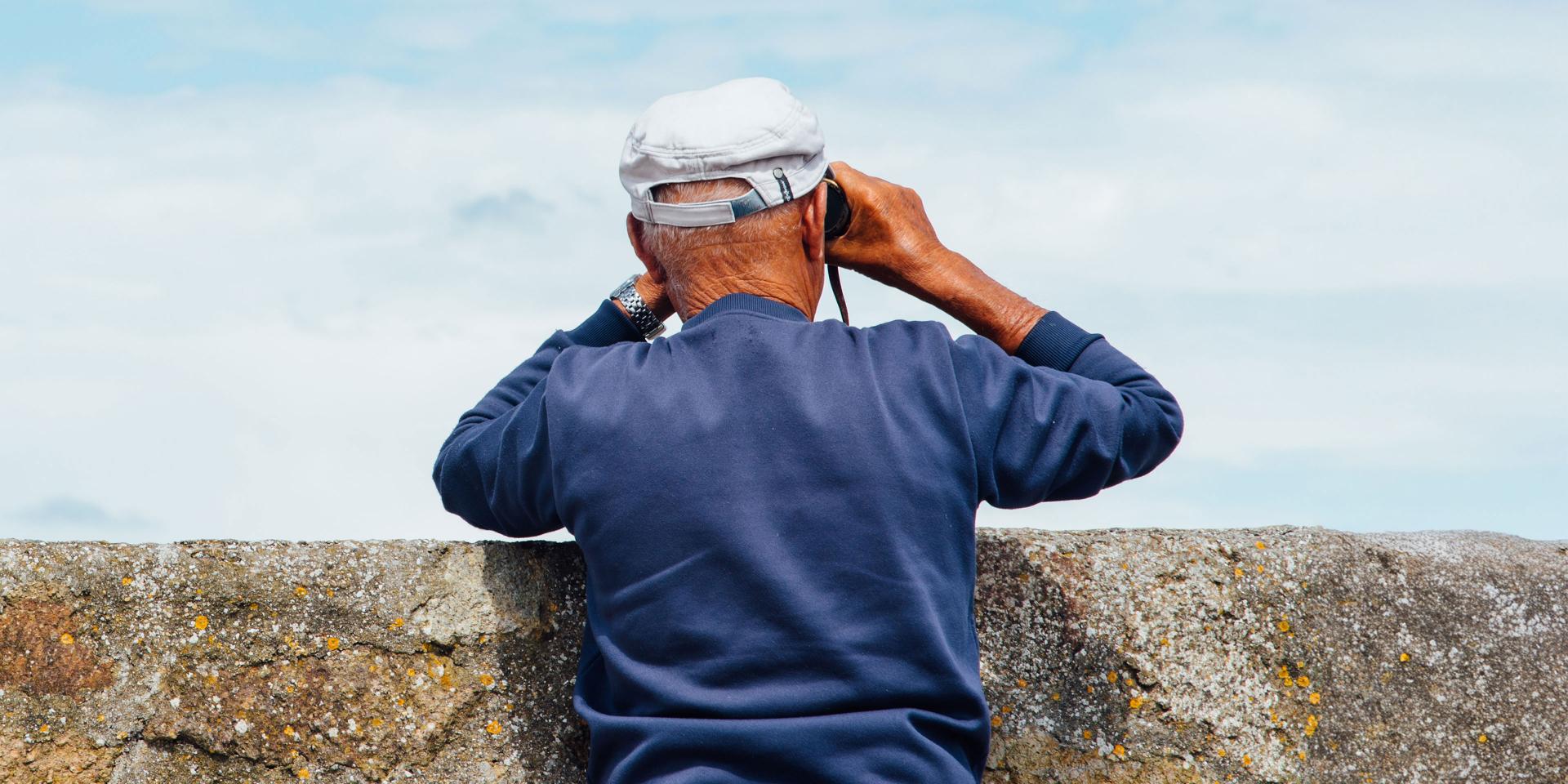 An older man wearing a blue sweater and white cap looks through binoculars over a stone wall toward the sky.