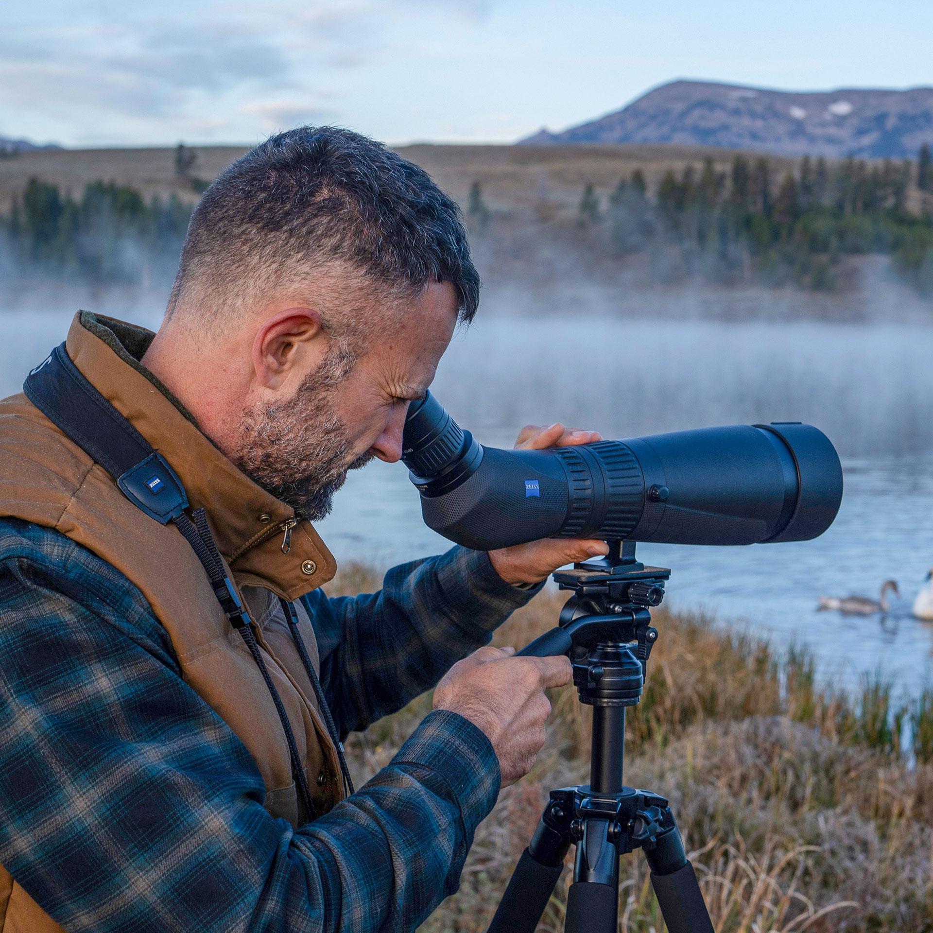 Man using a ZEISS spotting scope mounted on a tripod to observe wildlife by a misty lakeside.