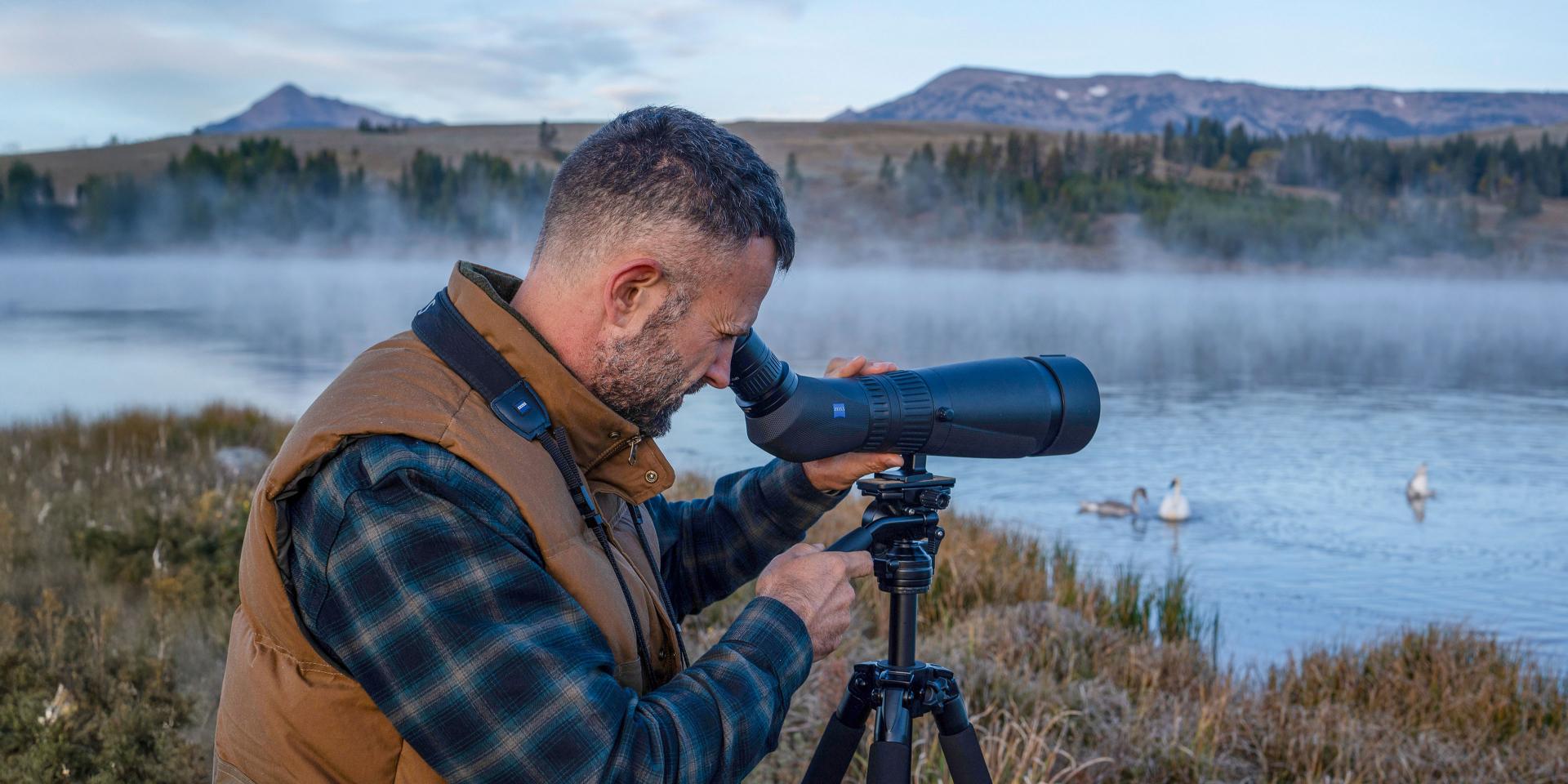 Man using a ZEISS spotting scope mounted on a tripod to observe wildlife by a misty lakeside.