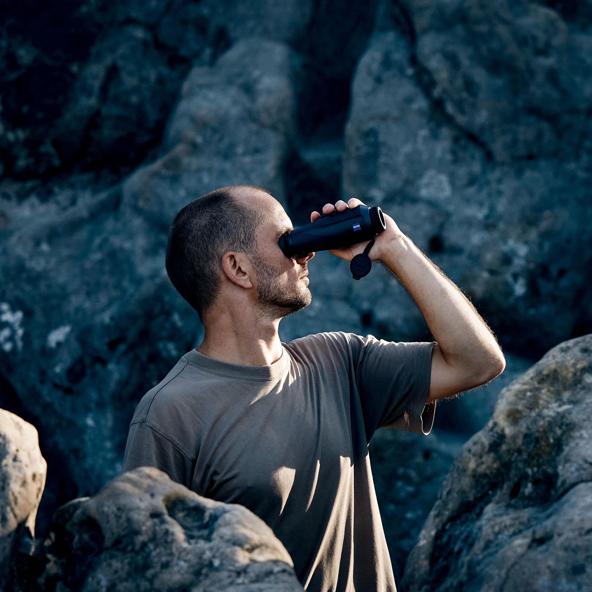 A man standing among large rocks looks through a pair of ZEISS binoculars.
