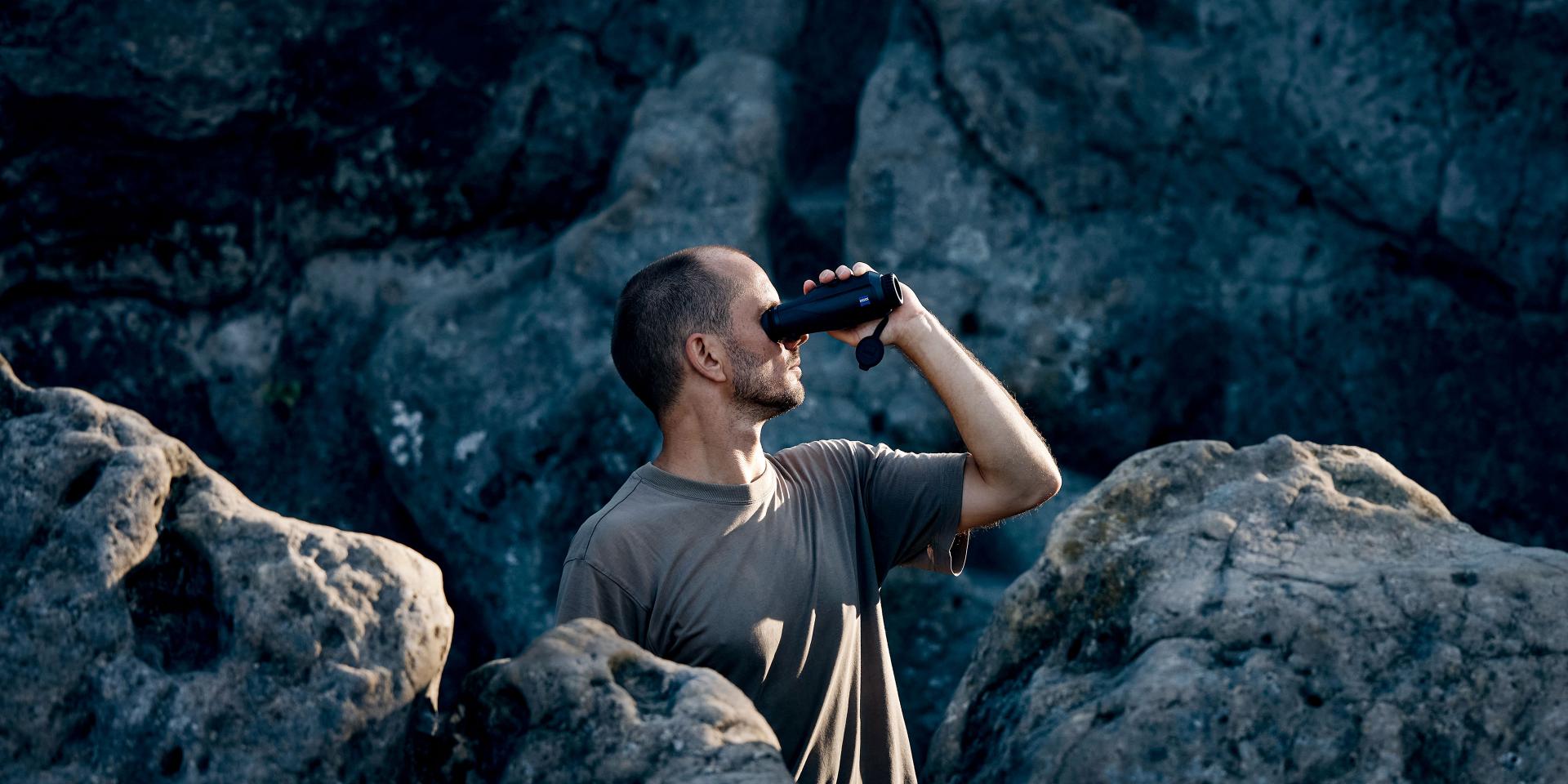 A man standing among large rocks looks through a pair of ZEISS binoculars.