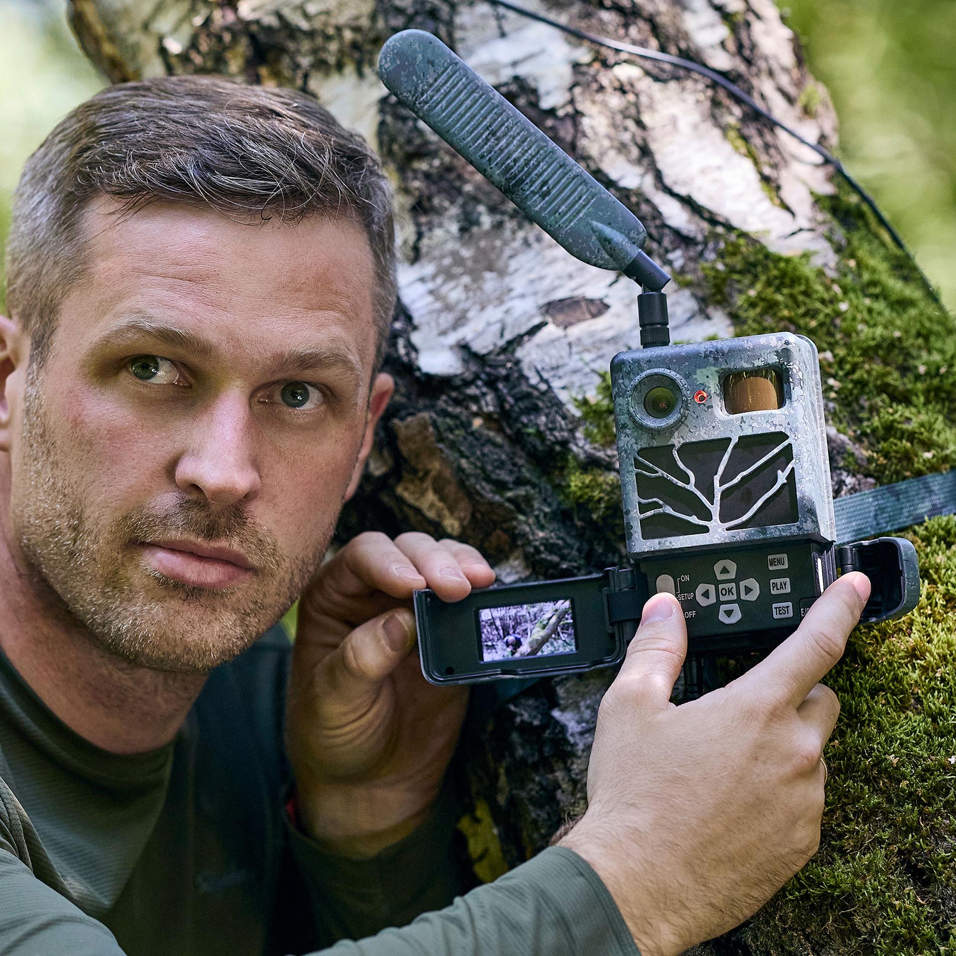 A man in outdoor clothing is using a ZEISS device while checking a trail camera mounted on a moss-covered tree in a forest.