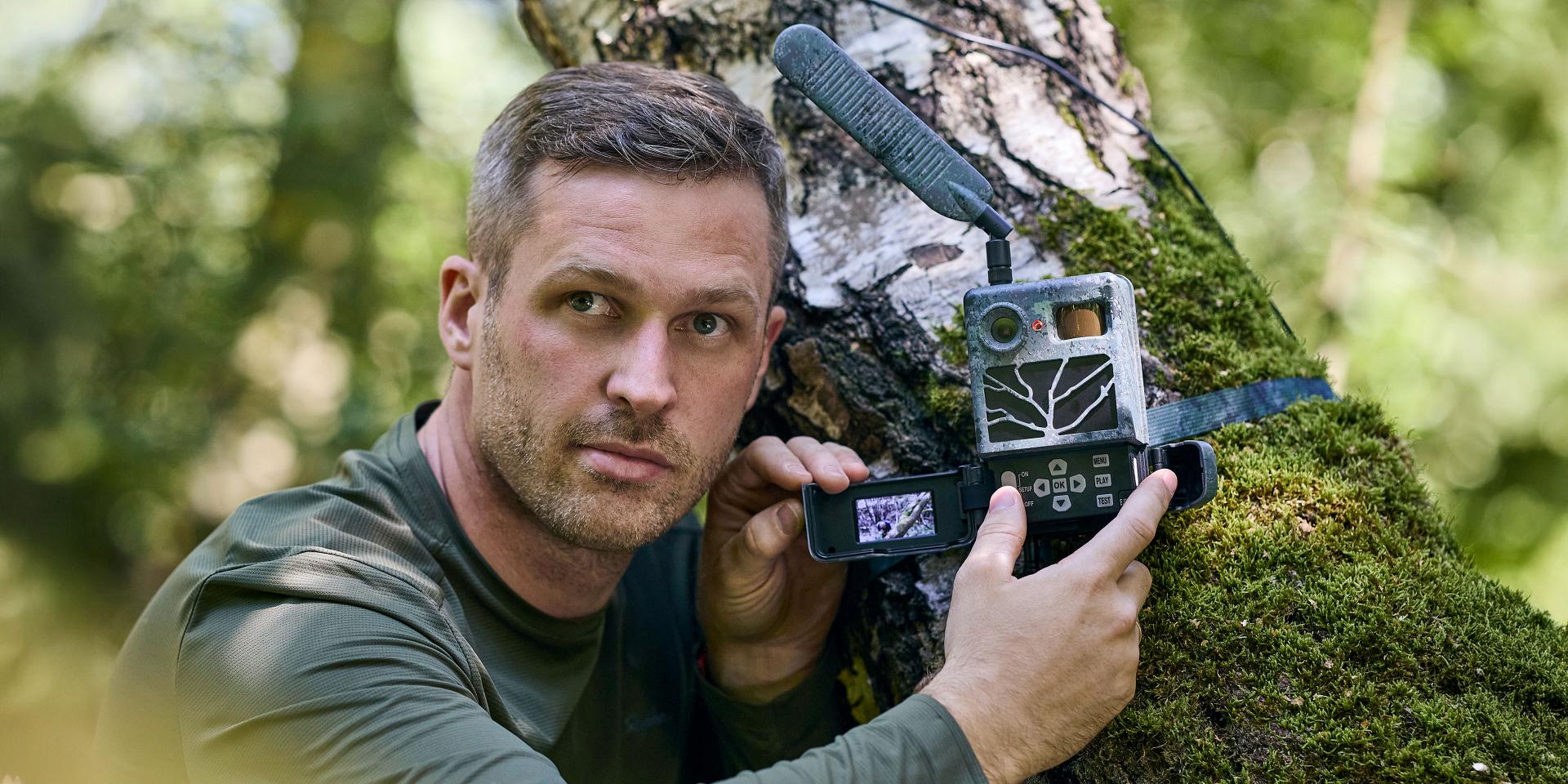 A man in outdoor clothing is using a ZEISS device while checking a trail camera mounted on a moss-covered tree in a forest.