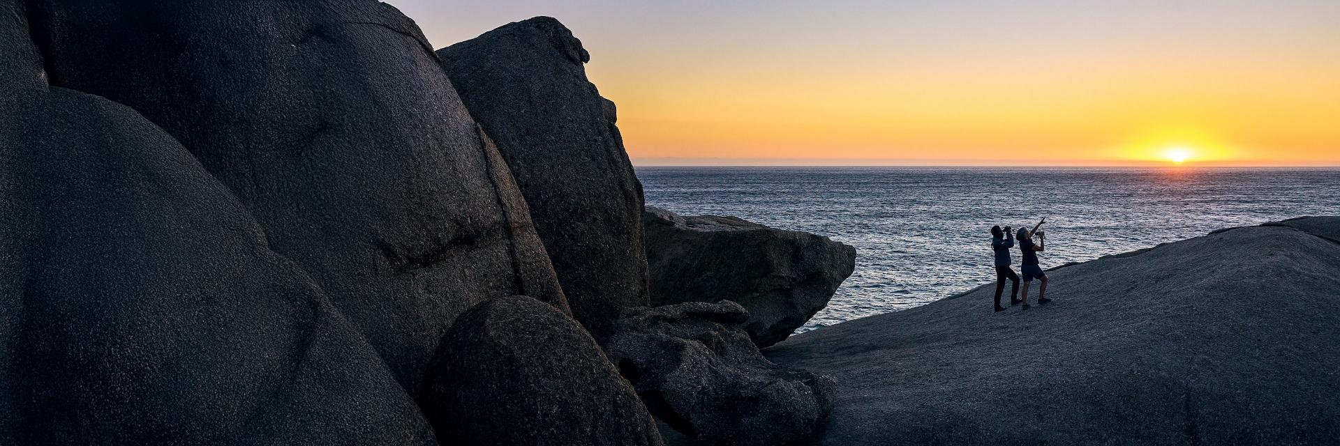 Two people standing on a large rocky shoreline at sunset, overlooking the ocean.