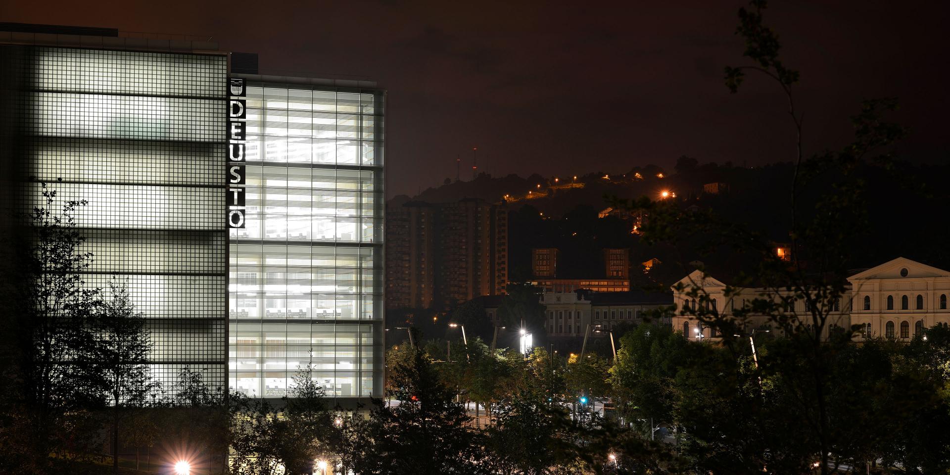 Modern glass building labeled "DEUSTO" illuminated at night, with surrounding city lights and hillside in the background.