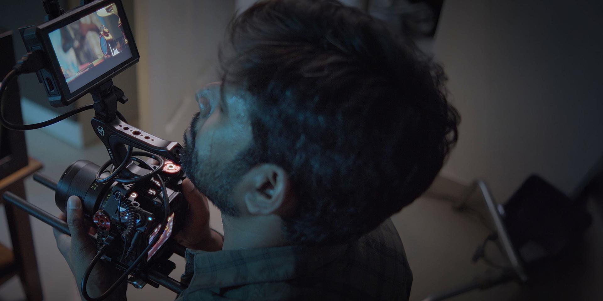 A videographer kneels on outdoor steps filming a person standing in white sneakers using a professional camera rig, with strong sunlight casting shadows.
