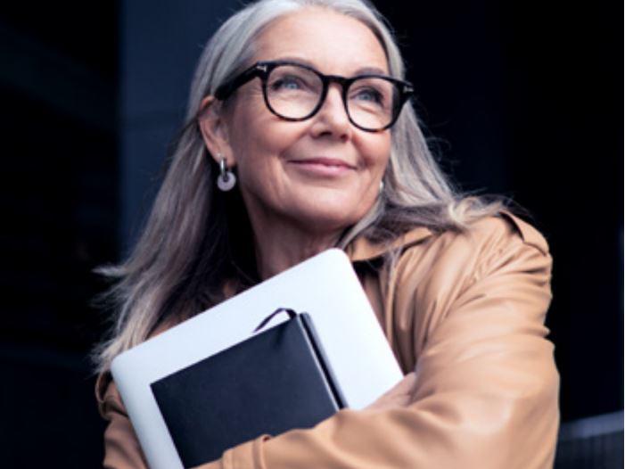 Una foto de una mujer mayor con gafas que sonríe y lleva un portátil.