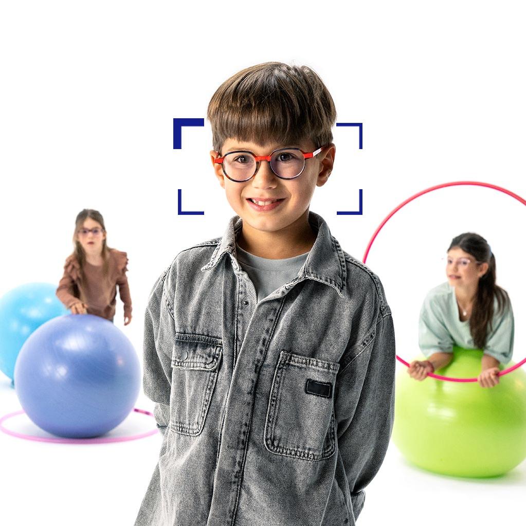 Un niño con gafas redondas de lentes ZEISS MyoCare se encuentra en primer plano y sonríe a la cámara. Al fondo, dos niñas con lentes ZEISS MyoCare jugando con aros y pelotas de gimnasia.