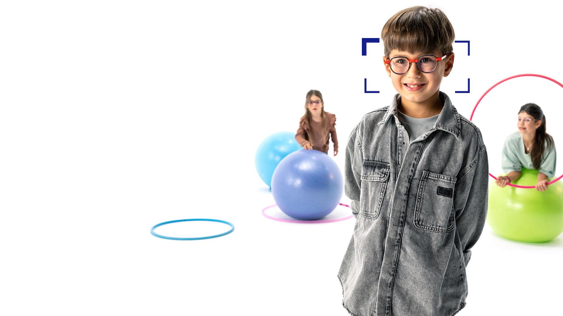 Un niño con gafas redondas de lentes ZEISS MyoCare se encuentra en primer plano y sonríe a la cámara. Al fondo, dos niñas con lentes ZEISS MyoCare jugando con aros y pelotas de gimnasia.