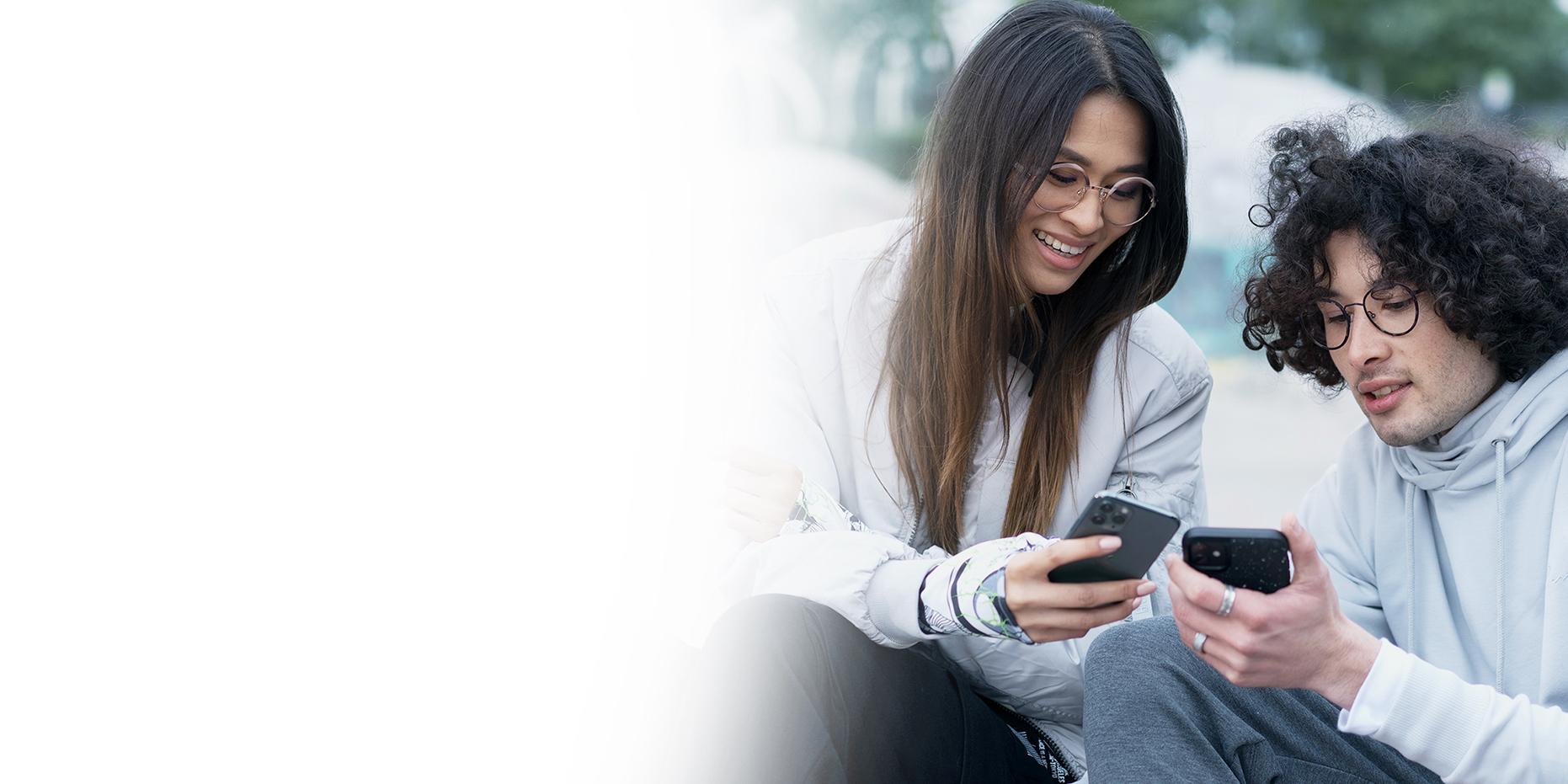 Dos jóvenes, una joven con pelo largo castaño sonriendo y un joven de pelo negro rizado con unas gafas ZEISS SmartLife, consultan sus teléfonos. 