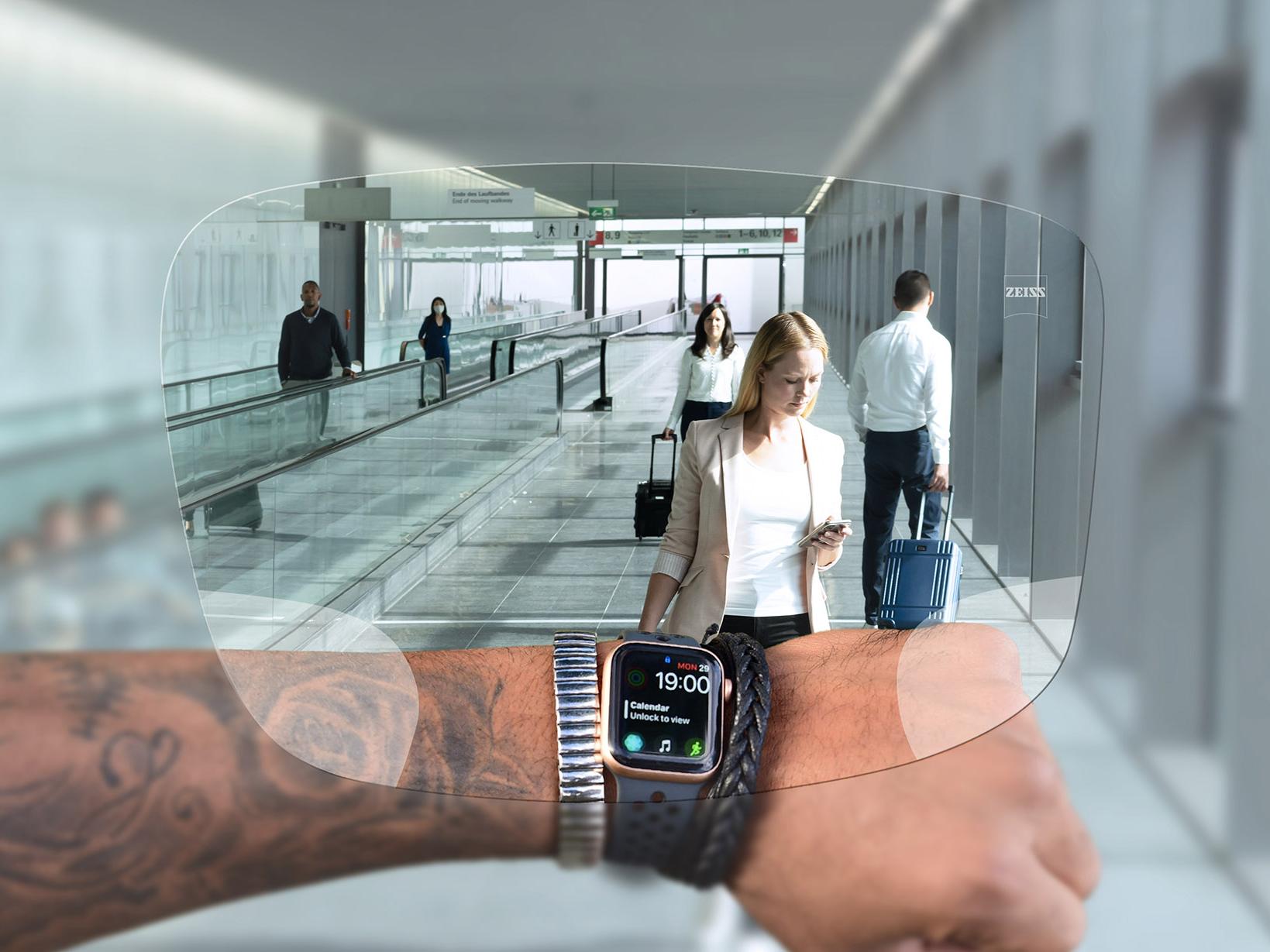Una persona consulta su reloj a través de las lentes ZEISS Digital SmartLife. Escena típica de un aeropuerto con personas caminando en cintas mecánicas empujando sus maletas. 