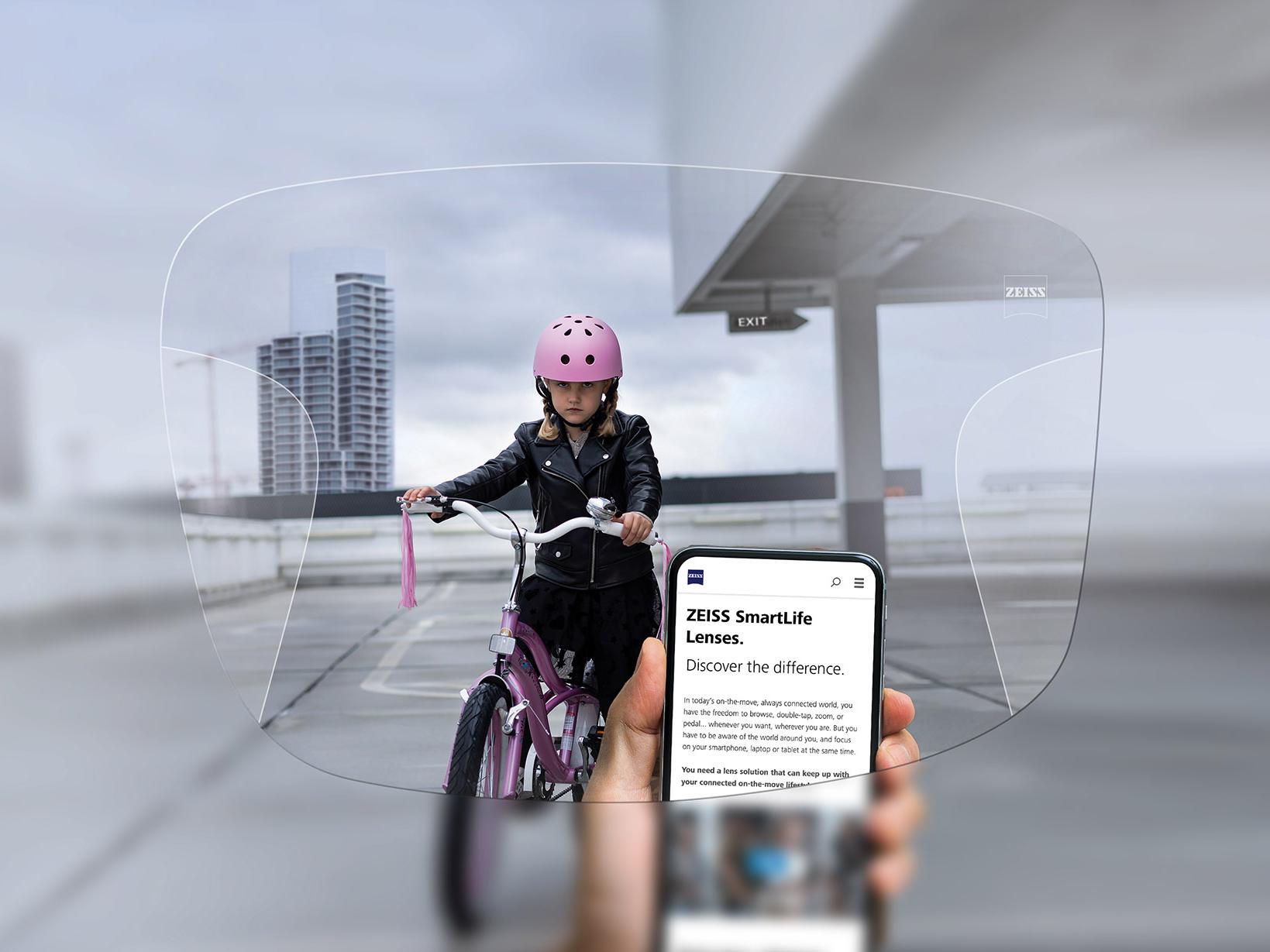 Una persona consulta su teléfono a través de las lentes progresivas ZEISS SmartLife. De fondo se puede ver a una niña con un casco rosa en una bicicleta. 