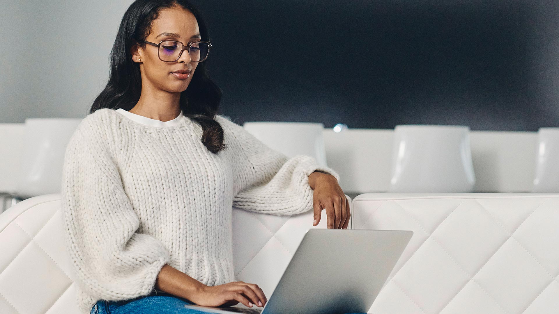 Una mujer de cabello oscuro, gafas oscuras y suéter blanco utiliza un portátil.
