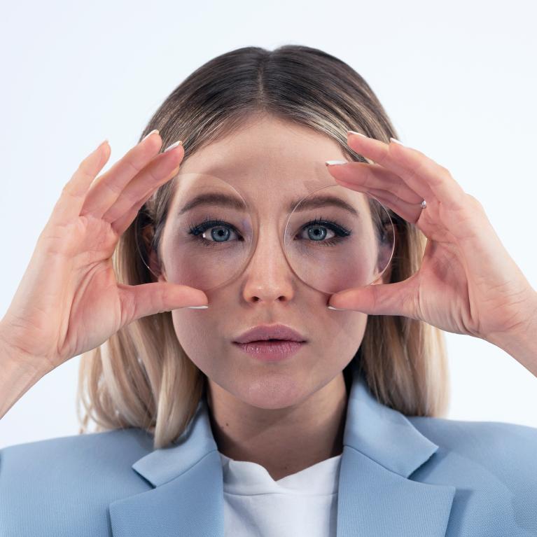 Una joven rubia sostiene unas lentes delante de los ojos para mostrar un buen aspecto sin ningún efecto raro gracias a las lentes monofocales ZEISS ClearView.