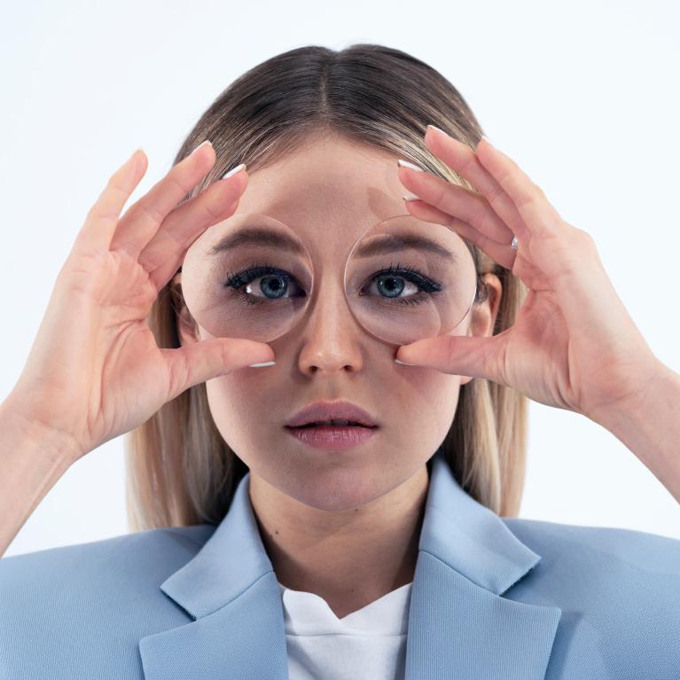 Una joven rubia sostiene unas lentes delante de los ojos para mostrar el efecto de ojo de pez causado por unas gafas gruesas.