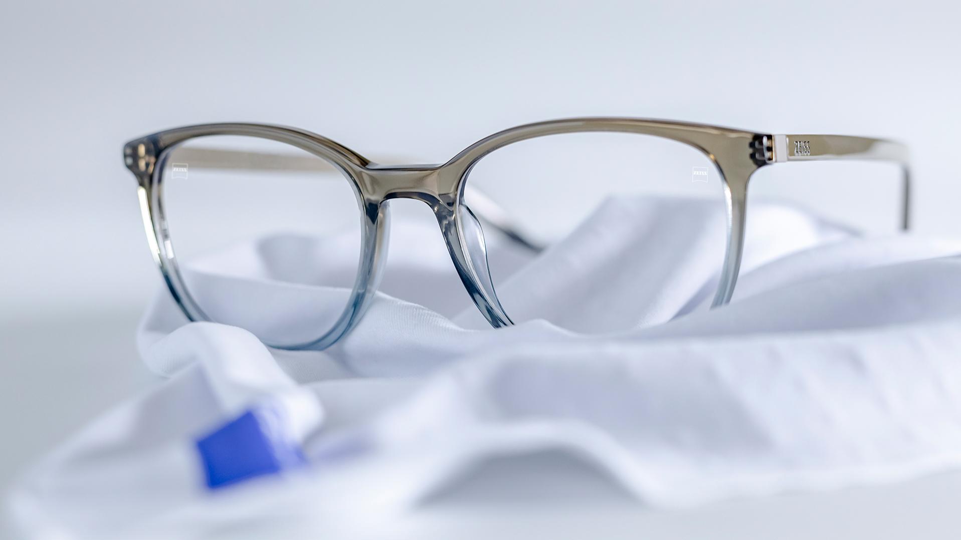 Un par de gafas con montura gris-azul y lentes ZEISS con protector DuraVision® reposan sobre un paño de microfibra blanco.
