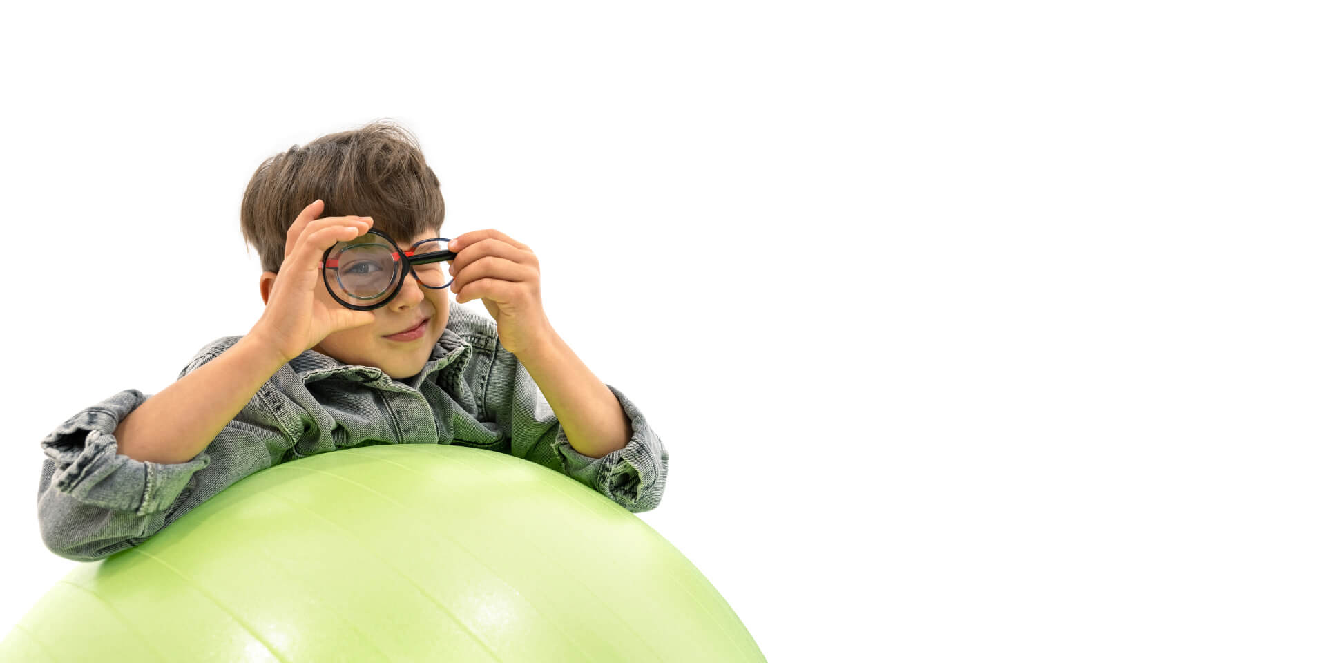 Un niño con lentes para el control de la miopía ZEISS apoyado en una pelota de gimnasia y sosteniendo una lupa delante de un ojo.