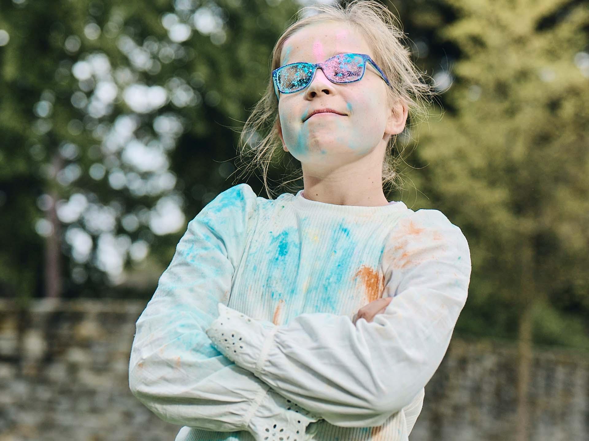 Una niña de brazos cruzados y con las gafas sucias de jugar con polvos de colores mira con descaro y sonríe.